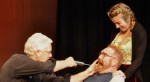 Actor injecting the mouth of another while a nurse holds him down